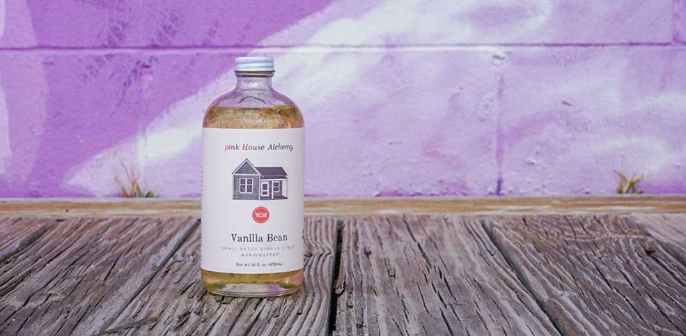 pink House Alchemy syrup bottle photographed in front of the purple wall in Seaside, FL. It is their vanilla flavor syrup for coffee.